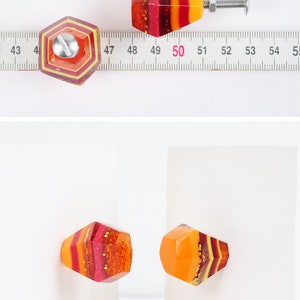 Op de afbeelding: Twee kleurrijke hars kastknoppen met een geometrische vorm en een gelaagd ontwerp in tinten rood, oranje en geel. De knoppen hebben een glanzende, glinsterende afwerking.