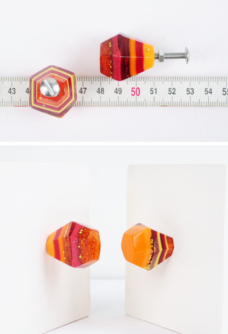 Op de afbeelding: Twee kleurrijke hars kastknoppen met een geometrische vorm en een gelaagd ontwerp in tinten rood, oranje en geel. De knoppen hebben een glanzende, glinsterende afwerking.