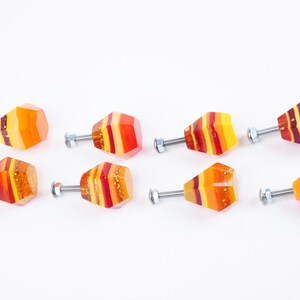 Op de afbeelding: Acht kleurrijke glazen kastknoppen met een gelaagd ontwerp in tinten rood, oranje en geel. Elke knop heeft een zilveren schroef.