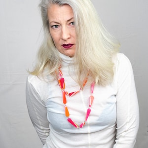 Könnte beinhalten: Eine lange Halskette mit geometrischen Formen in Orange- und Rosatönen. Die Kette wird über einem weißen Rollkragenpullover getragen. Die Formen sind durch silberne Glieder verbunden. Die Frau hat graues Haar und dunklen Lippenstift.