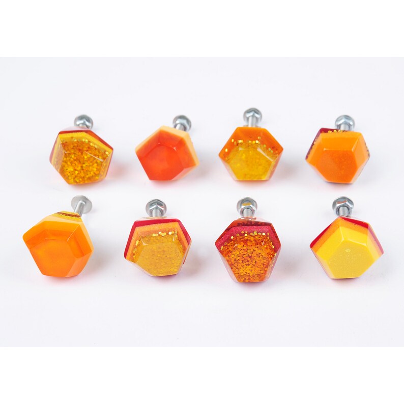 Op de afbeelding: Acht kleurrijke zeshoekige kastknoppen met een metalen afwerking. De knoppen zijn gelaagd met tinten rood, oranje en geel met glitteraccenten.