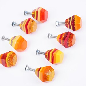 Op de afbeelding: Acht kleurrijke kastknoppen met een geometrische vorm. De knoppen zijn gemaakt van hars en hebben een gelaagd ontwerp met tinten rood, oranje en geel. Elke knop heeft een zilveren schroef.