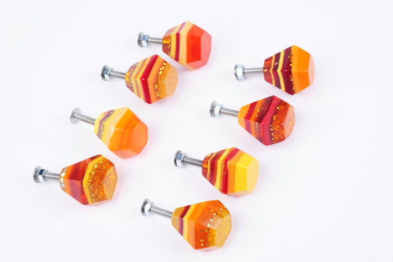 Op de afbeelding: Acht kleurrijke kastknoppen met een geometrische vorm. De knoppen zijn gemaakt van hars en hebben een gelaagd ontwerp met tinten rood, oranje en geel. Elke knop heeft een zilveren schroef.