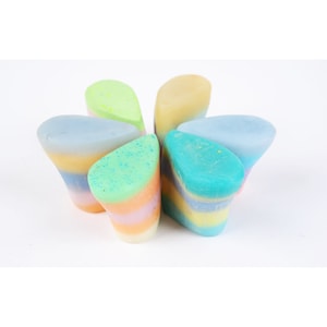 Op de afbeelding: Een verzameling van zes kleurrijke, gelaagde zepen, gerangschikt in een cirkelvormig patroon. Elke zeep heeft een unieke kleurencombinatie, waaronder blauw, groen, geel en oranje, met een gespikkelde bovenkant. De zepen staan op een witte achtergrond.