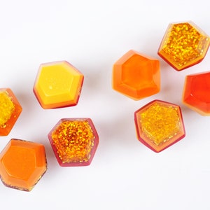 Op de afbeelding: Acht zeshoekige hars cabochons in tinten van oranje en geel met gouden glitter. De cabochons zijn verspreid op een witte achtergrond.