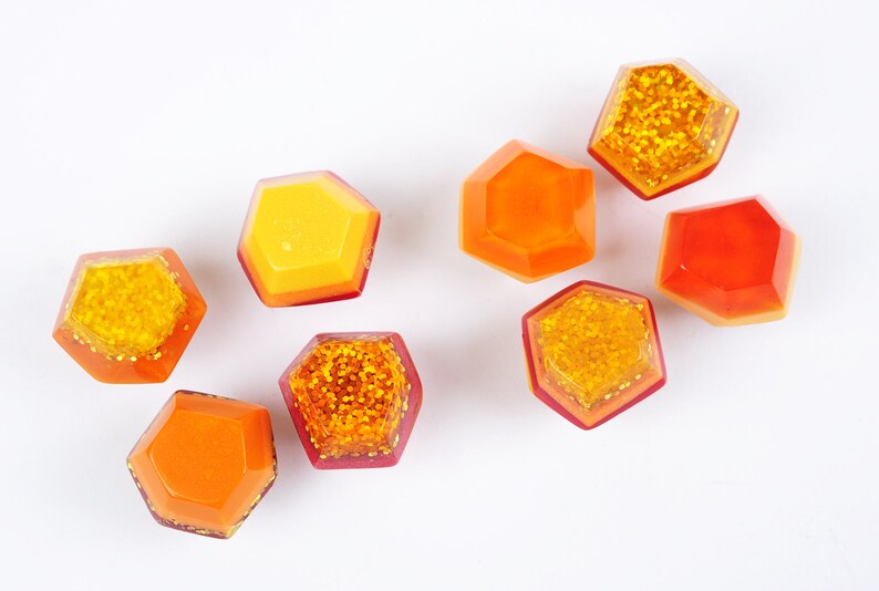 Op de afbeelding: Acht zeshoekige hars cabochons in tinten van oranje en geel met gouden glitter. De cabochons zijn verspreid op een witte achtergrond.