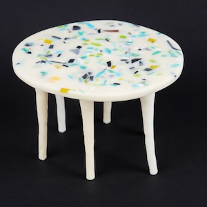 Puede incluir: Una mesa redonda pequeña y blanca con una tapa blanca y cinco patas blancas. La tapa está cubierta de piezas de vidrio coloridas, como confeti.