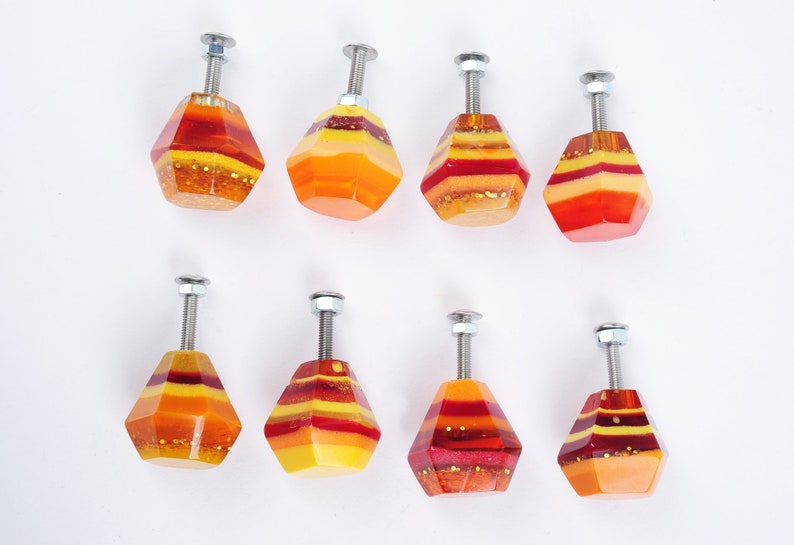 Op de afbeelding: Acht kleurrijke, geometrische kastknoppen met een gelaagd ontwerp in tinten rood, oranje en geel. Elke knop heeft een zilveren metalen schroef.