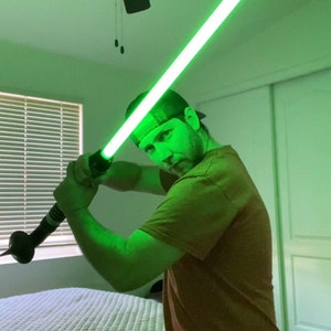 May include: A person holding a green lightsaber in a bedroom with a ceiling fan. The lightsaber is lit up and the person is wearing a brown shirt and a baseball cap.