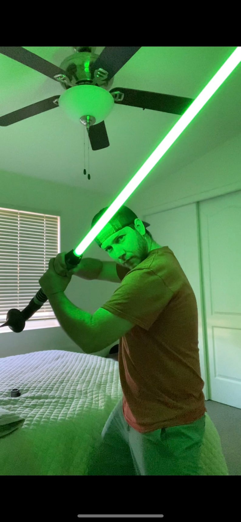 May include: A person holding a green lightsaber in a bedroom with a ceiling fan. The lightsaber is lit up and the person is wearing a brown shirt and a baseball cap.