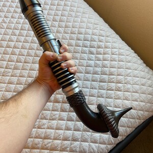 May include: A silver and black lightsaber with a curved, brown handle. The handle has a textured, ring-like design at the end.