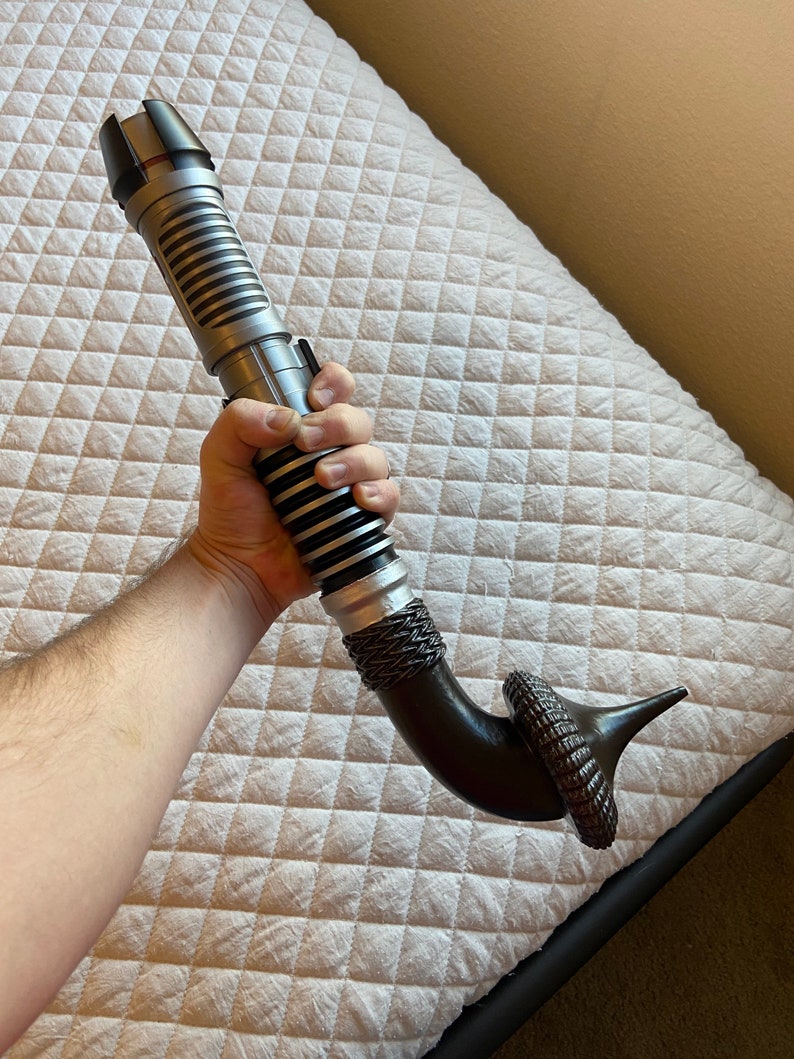 May include: A silver and black lightsaber with a curved, brown handle. The handle has a textured, ring-like design at the end.