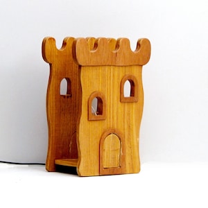 Puede incluir: Un castillo de madera de juguete con un acabado marrón. El castillo tiene un techo almenado, tres ventanas y una puerta.