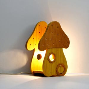 Puede incluir: Una lámpara de madera con forma de hongo con un acabado marrón claro. La lámpara tiene una pequeña bombilla en el interior, iluminando el interior del hongo.