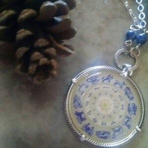 May include: Silver pendant necklace with a circular zodiac design in blue. The pendant hangs from a silver chain with blue beads. A brown pine cone is in the background.