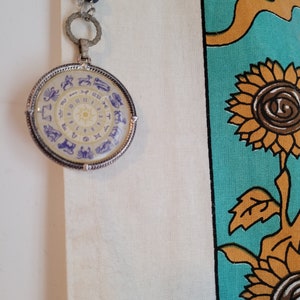 Puede incluir: Un collar de plata con un colgante redondo que presenta una rueda zodiacal blanca y amarilla con símbolos astrológicos morados. El colgante cuelga sobre un fondo de tela blanca con un patrón floral azul y amarillo.