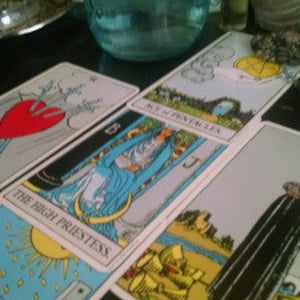 May include: A blue glass jar with a silver lid is positioned near several tarot cards. The cards display colourful illustrations and text, including "The High Priestess" and "Ace of Pentacles." The image evokes a mystical or spiritual atmosphere.