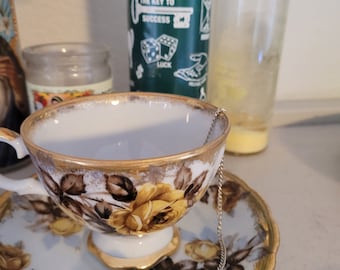 Könnte beinhalten: Ein Vintage-Tassen- und Untertassen-Set mit einem Blumenmuster in Gelb und Braun, akzentuiert mit goldenen Verzierungen. Ein grünes Kristallpendel hängt über der Tasse. Eine grüne Kerze mit Text befindet sich im Hintergrund.