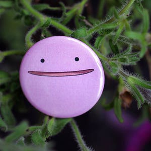 May include: A pink button with a cartoon face. The face has two black eyes and a wide, smiling mouth.