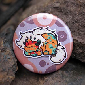 May include: A round button with a colorful cartoon animal on it. The animal is a white, orange, and green creature with a red mouth and black eyes. The button has a pink, purple, and white background with a pattern of circles.