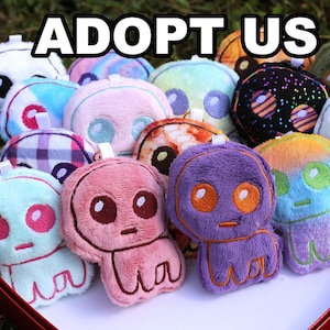 May include: A selection of colourful plush alien toys, including pink, purple, and blue, presented in a heart-shaped box. The text "ADOPT US" is visible above the toys.
