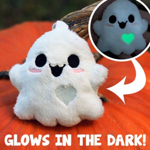 May include: A white plush ghost keychain with black eyes, a smiling mouth, and pink cheeks. The ghost has a heart-shaped cutout and glows green in the dark. The text "GLOWS IN THE DARK!" is at the bottom.