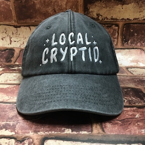 May include: A black baseball cap with white embroidered text that reads "*LOCAL* CRYPTID*"