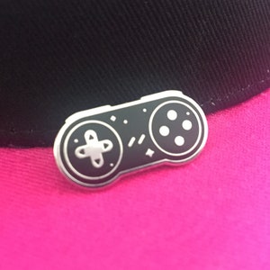 May include: A silver-coloured pin shaped like a classic video game controller. The controller is black with white buttons and a white directional pad. The pin is on a pink surface.