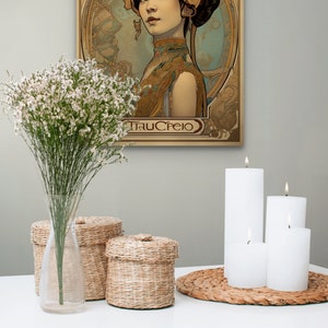 May include: A vintage-style poster featuring a woman with dark hair and a floral headdress. The poster is in a gold frame and has a light blue background. The text "TRUCREIO" is at the bottom of the poster. The poster is hanging on a wall with white candles and wicker baskets on a white table in front of it.
