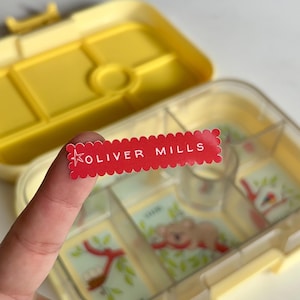 May include: A rectangular red sticker with a scalloped edge featuring a white star icon and the text OLIVER MILLS in white capital letters. The sticker is held in front of an open yellow plastic bento box with multiple compartments and animal graphics.