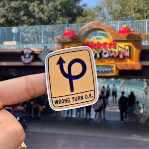 May include: A rectangular sticker with a light tan background and a dark blue graphic of a looping arrow. The text WRONG TURN O.K. is printed in dark blue capital letters at the bottom of the sticker, which has a white border.
