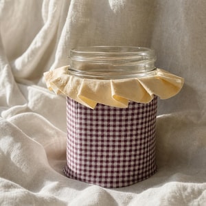 May include: A clear glass jar wrapped in a dark maroon and white gingham fabric sleeve. The top of the jar features a decorative, ruffled pale yellow fabric trim that encircles the rim, creating a layered and textured appearance on the container.