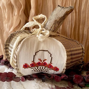 May include: A fabric pumpkin decoration featuring brown and white gingham side panels and a central white panel with a cross-stitched basket of red and purple flowers. The pumpkin is topped with a natural wooden stem and a cream-colored ribbon.