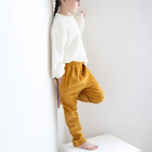 May include: A child in a cream ribbed knit sweater and mustard yellow corduroy trousers. The trousers have an elasticated waistband and a relaxed fit. The outfit is casual and comfortable.