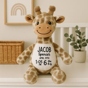 Op de afbeelding: Een zacht girafspeelgoed met een lachend gezicht en bruine en bruine vlekken. De giraf heeft de naam "Jacob Spencer" en geboortedetails op zijn buik: 29 april, 13:05 uur, 2,72 kg.