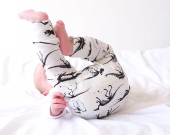 May include: A baby wearing white and black leggings with a dinosaur print. The baby is lying on its back on a white sheet.