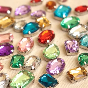 May include: Assortment of colorful, faceted glass rhinestones with silver settings. The rhinestones are in various shapes, including round, oval, pear, and square.