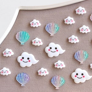 May include: A collection of white cloud and iridescent seashell shaped resin embellishments. The clouds have happy faces and pink cheeks.