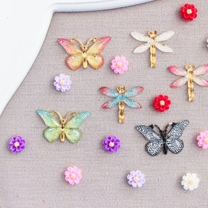May include: An assortment of decorative embellishments including butterflies, dragonflies, and small flowers in various colors. The butterflies and dragonflies have metallic gold accents. The flowers are in shades of pink, purple, red, and white.
