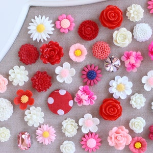 May include: Assortment of colourful flower-shaped buttons in red, pink, white, and blue. The buttons are arranged on a grey fabric background.