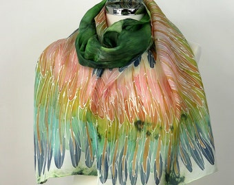 Puede incluir: Una bufanda de seda con un diseño pintado a mano de plumas verdes, naranjas y azules. La bufanda está drapeada sobre un maniquí blanco.