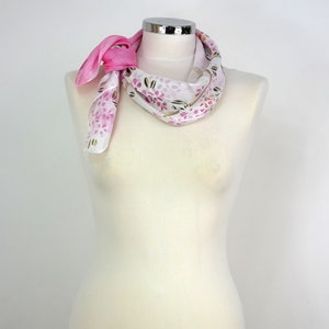 May include: A white silk scarf with a pink floral pattern and a pink trim. The scarf is tied around a white mannequin's neck.