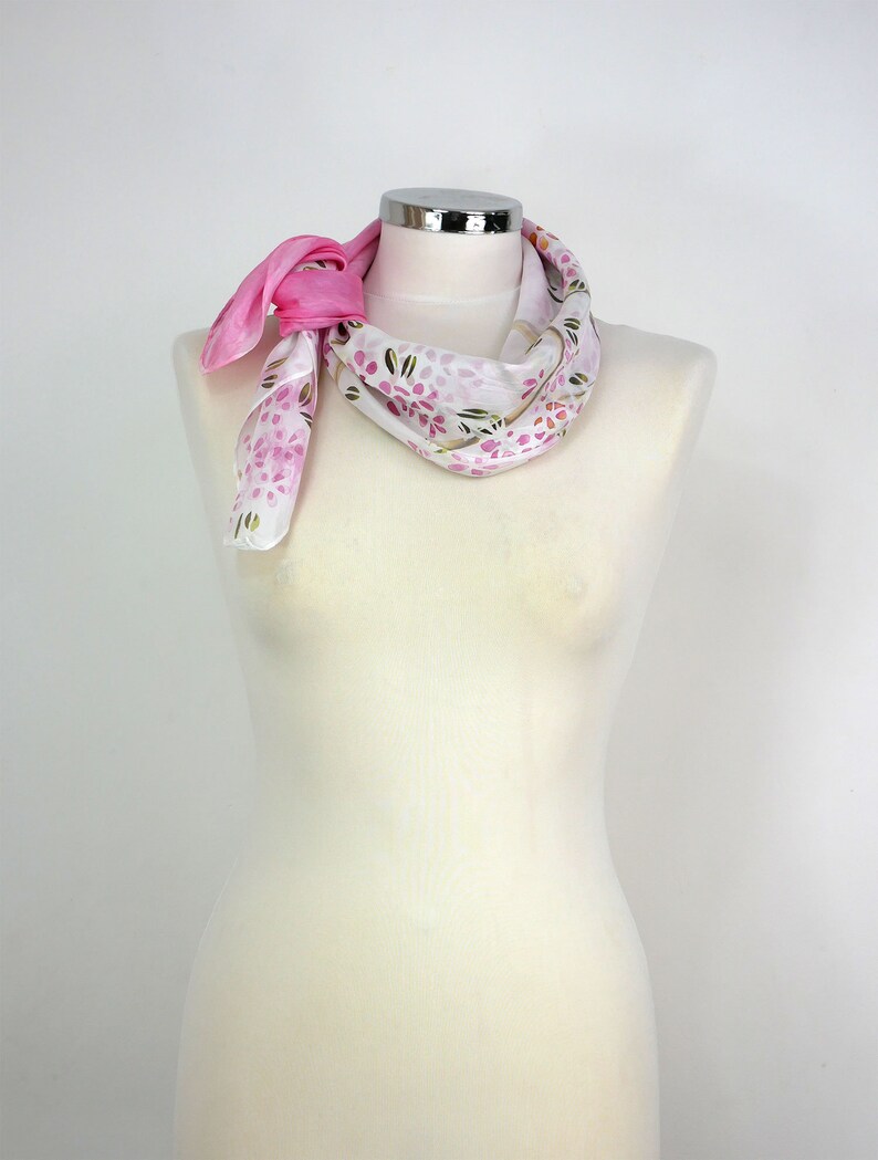 May include: A white silk scarf with a pink floral pattern and a pink trim. The scarf is tied around a white mannequin's neck.