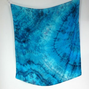 May include: A blue silk scarf with a tie-dye pattern. The scarf is hanging from a white background.