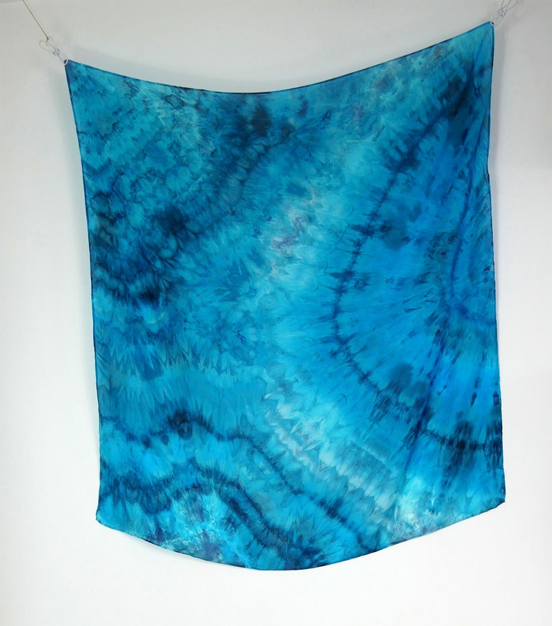 May include: A blue silk scarf with a tie-dye pattern. The scarf is hanging from a white background.