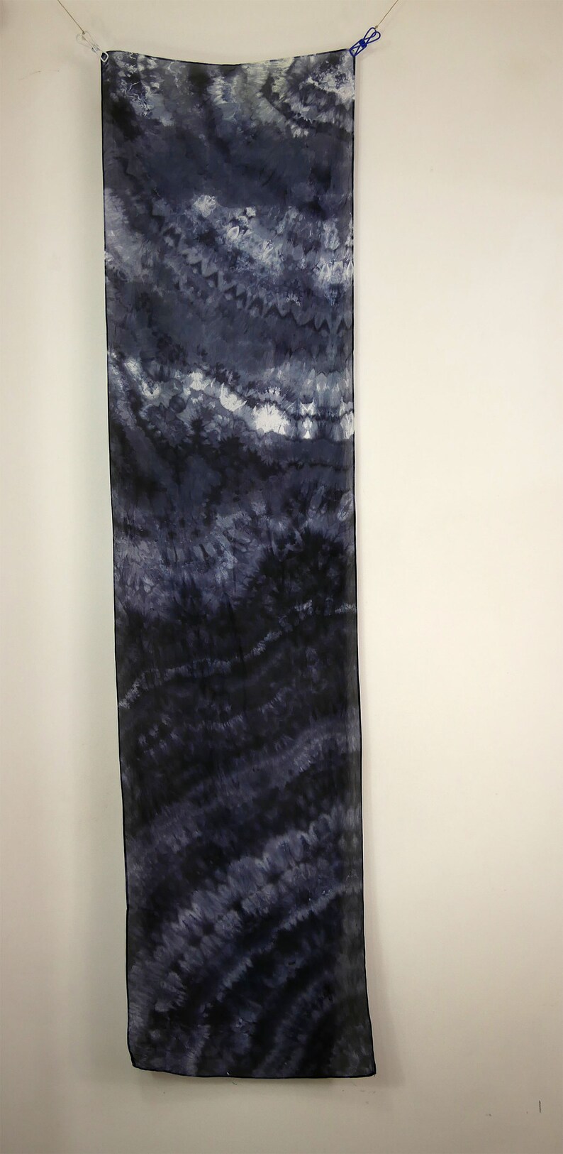 Op de afbeelding: Een lange, rechthoekige sjaal met een tie-dye patroon in tinten blauw en wit. De sjaal hangt aan een waslijn.