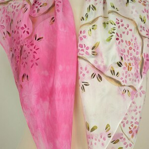 May include: A pink and white silk scarf with a floral pattern. The scarf is hand-painted with a design of branches and flowers.