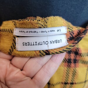 K&ouml;nnte beinhalten: Nahaufnahme eines gelb, orange und schwarz karierten Stoffes mit dem Etikett "URBAN OUTFITTERS" und "S-P Made in Turkey / Fabriqu&eacute; en Turquie". Der Stoff wird von einer Hand gehalten.
