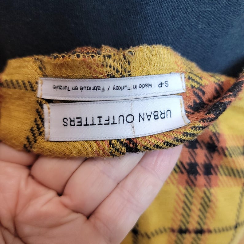 K&ouml;nnte beinhalten: Nahaufnahme eines gelb, orange und schwarz karierten Stoffes mit dem Etikett "URBAN OUTFITTERS" und "S-P Made in Turkey / Fabriqu&eacute; en Turquie". Der Stoff wird von einer Hand gehalten.