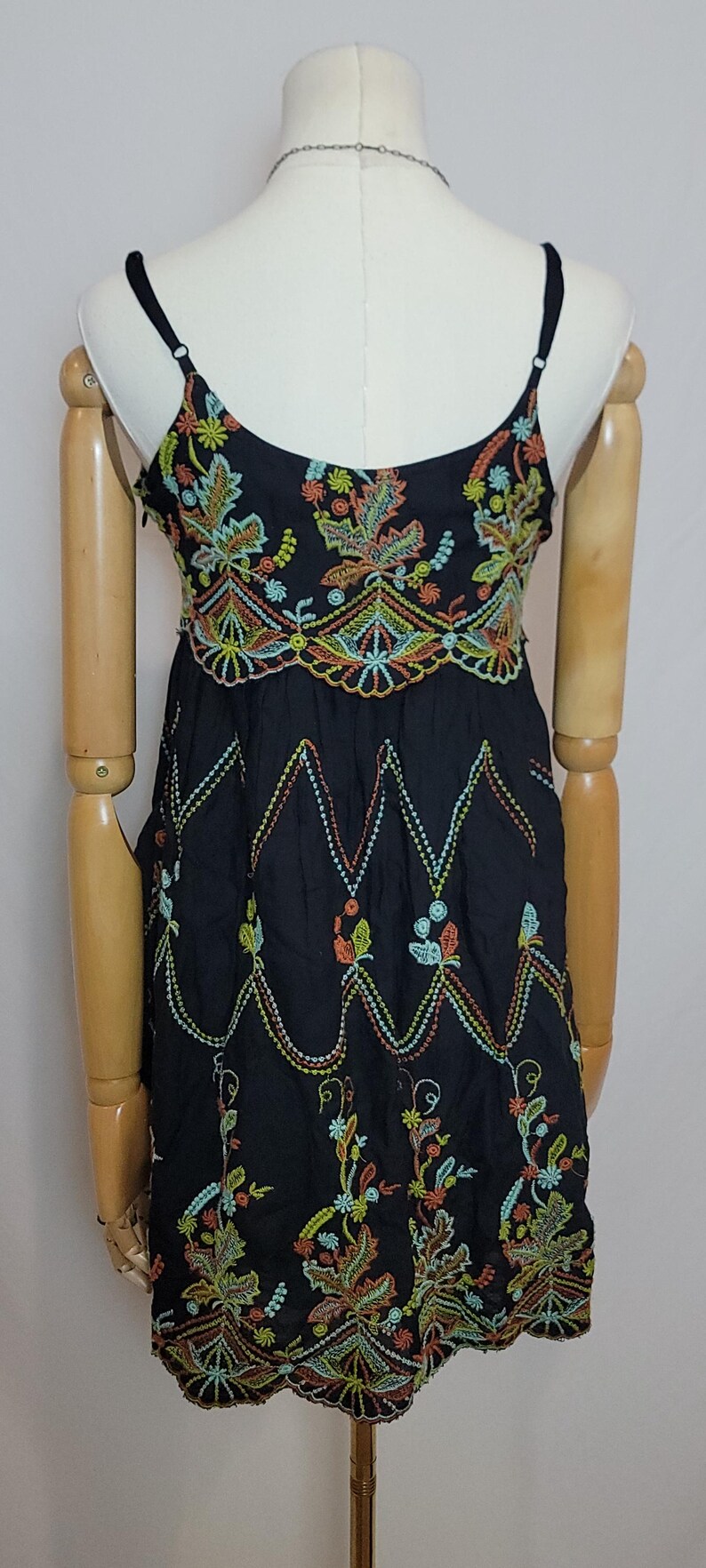 May include: Black spaghetti strap dress with colourful floral embroidery. The dress features intricate embroidery in shades of orange, green, yellow, and blue. The dress is displayed on a mannequin.